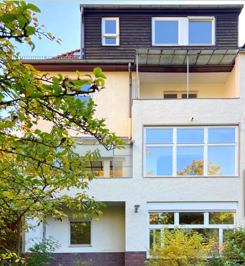 Die Gartenseite des Hauses mit großen Fenstern, Balkon und einem Obstbaum im Vordergrund