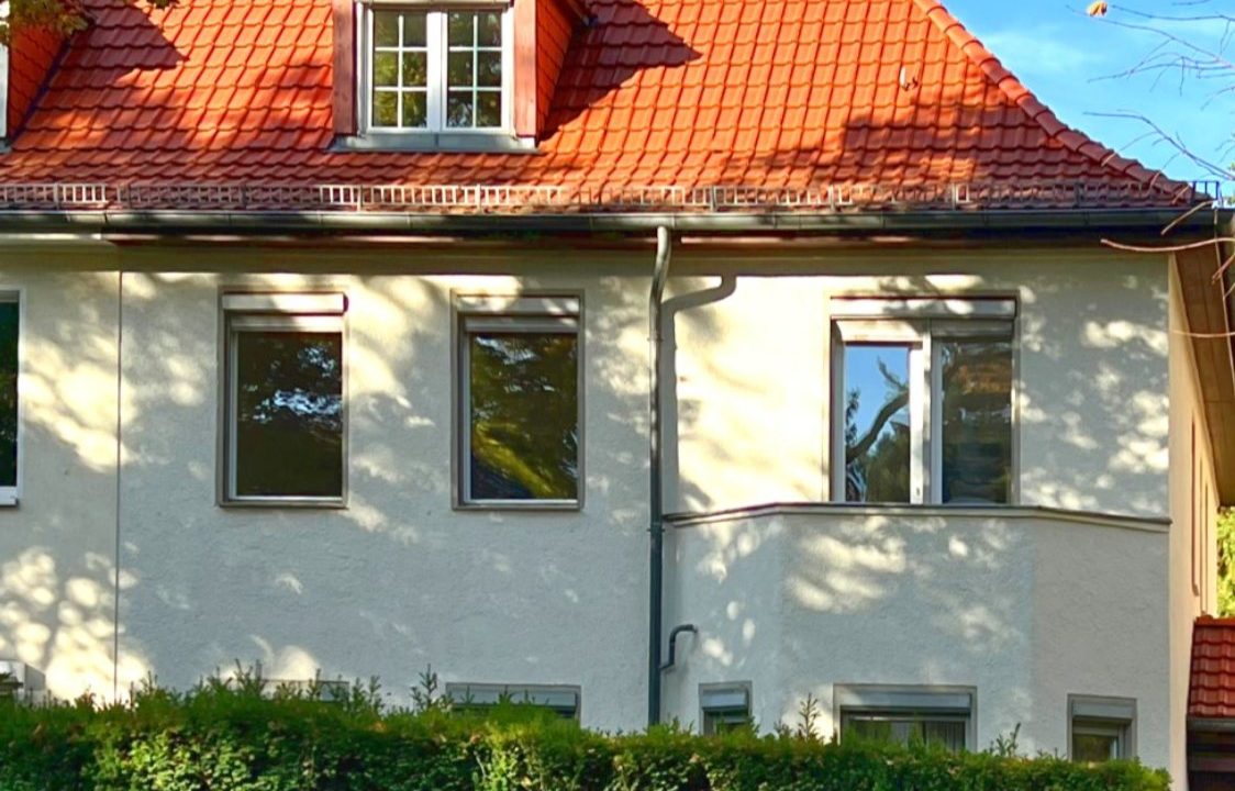 Die Doppelhaushälfte in der Grunewaldstraße 29 von der Straße aus: helle Putzfassade, rotes Ziegeldach mit Gaube, davor eine Hecke